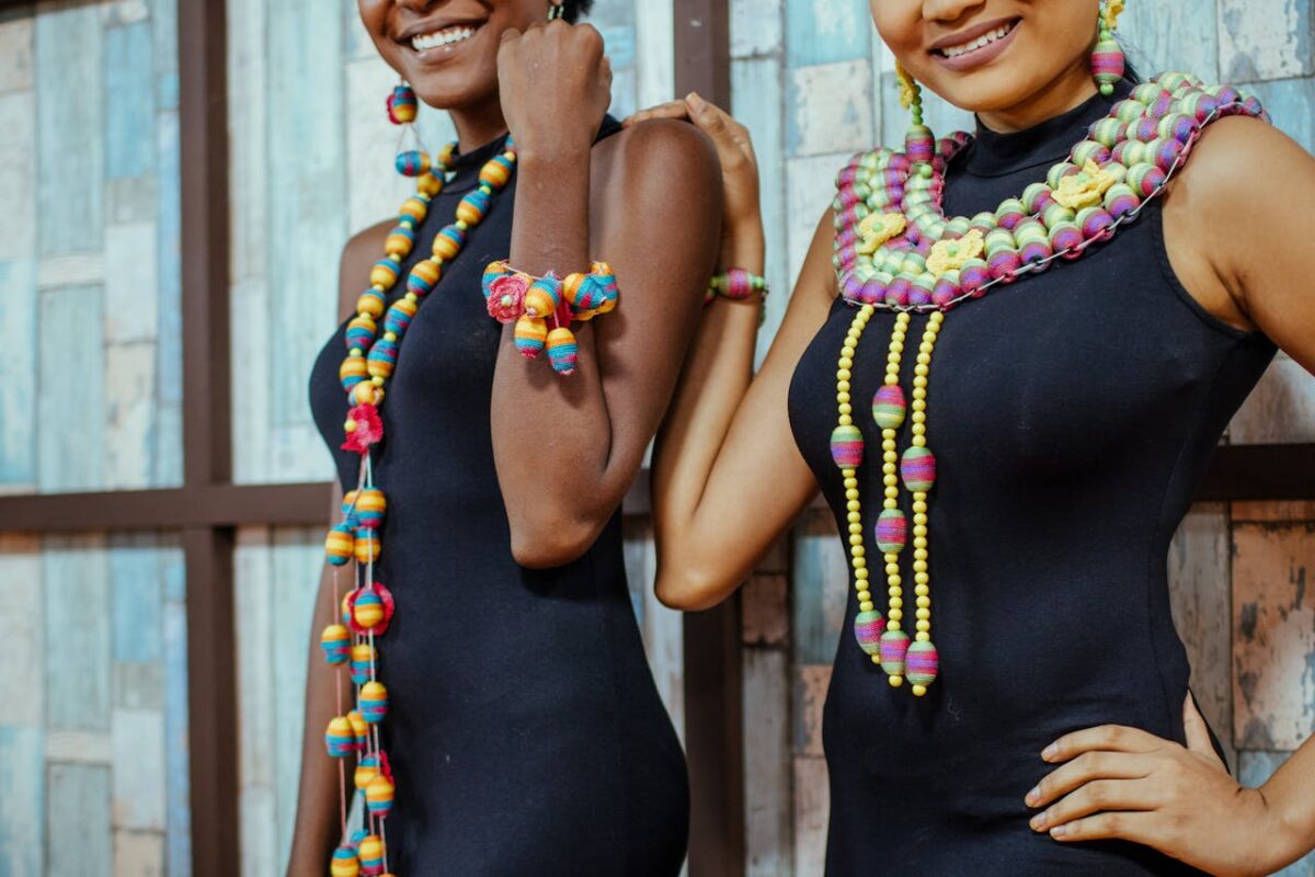Two African models showcasing vibrant beaded jewelry and elegant fashion against a rustic backdrop.