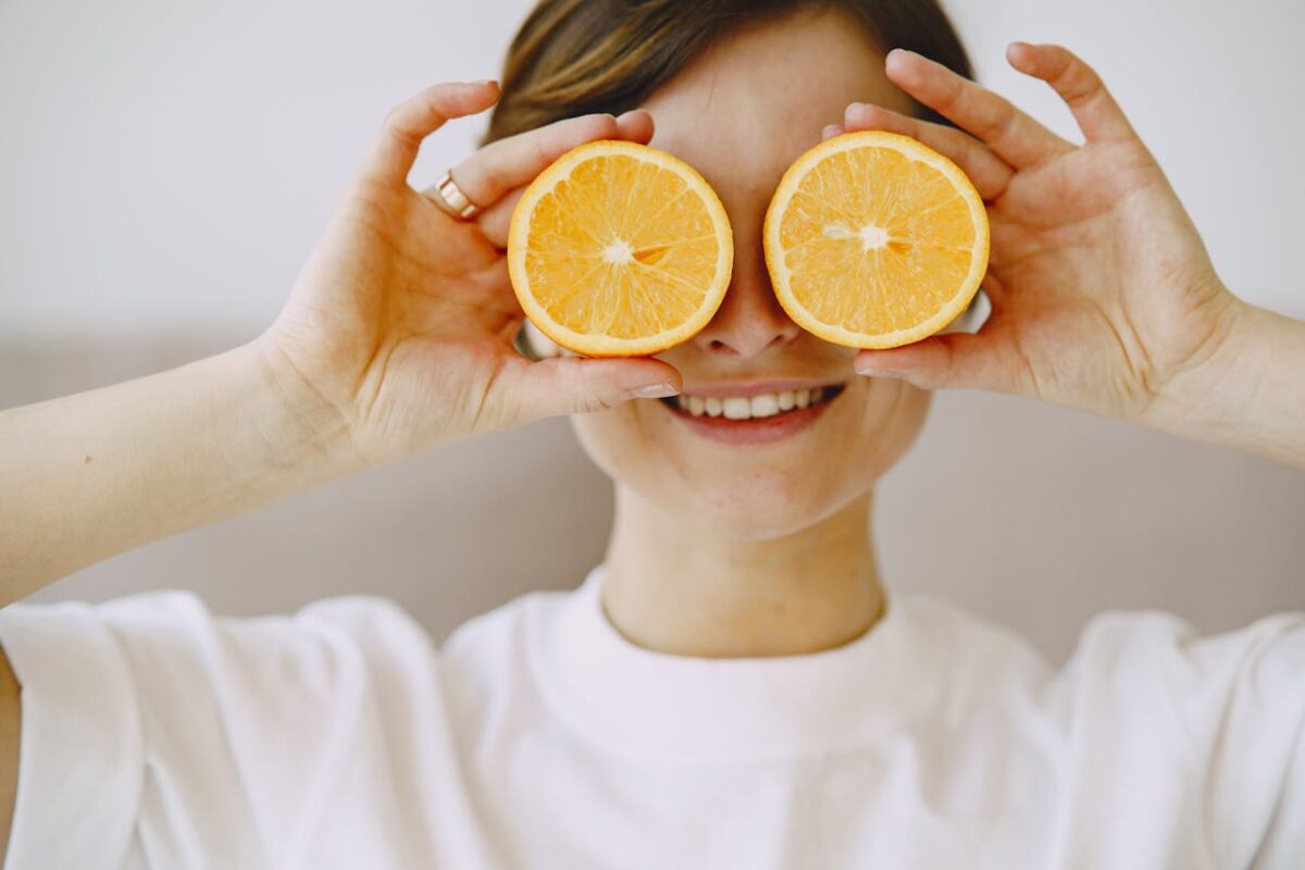 Crop positive female covering eyes with fresh slices of orange smiling and looking forward on blurred background